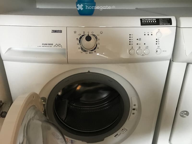 White washing machine with control panel, open door revealing interior, small blue container on top.