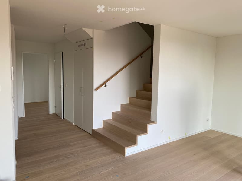 Empty hallway with wooden flooring, white walls, and a staircase leading upwards with a wooden handrail.