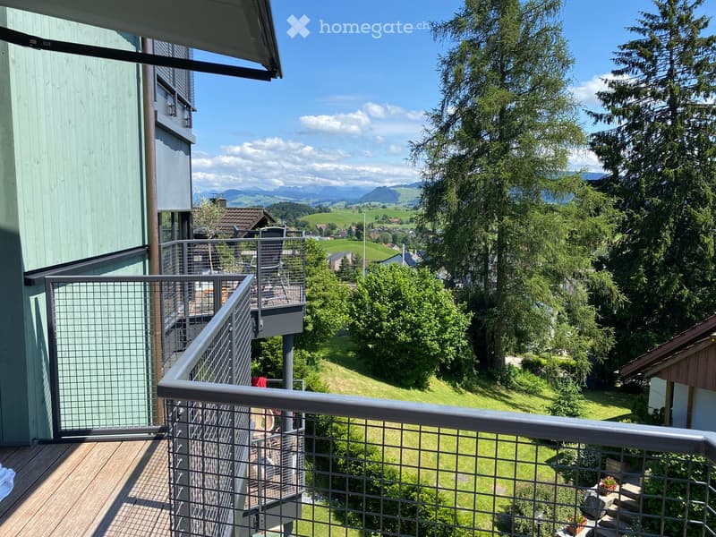 Balcony with metal railing, wooden decking, overlooking a green valley and distant mountains.