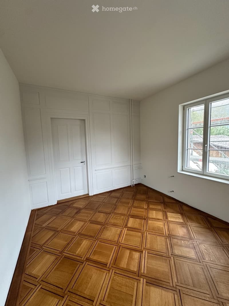 Empty room with wooden parquet flooring, white walls, and large windows. No furniture or equipment present.