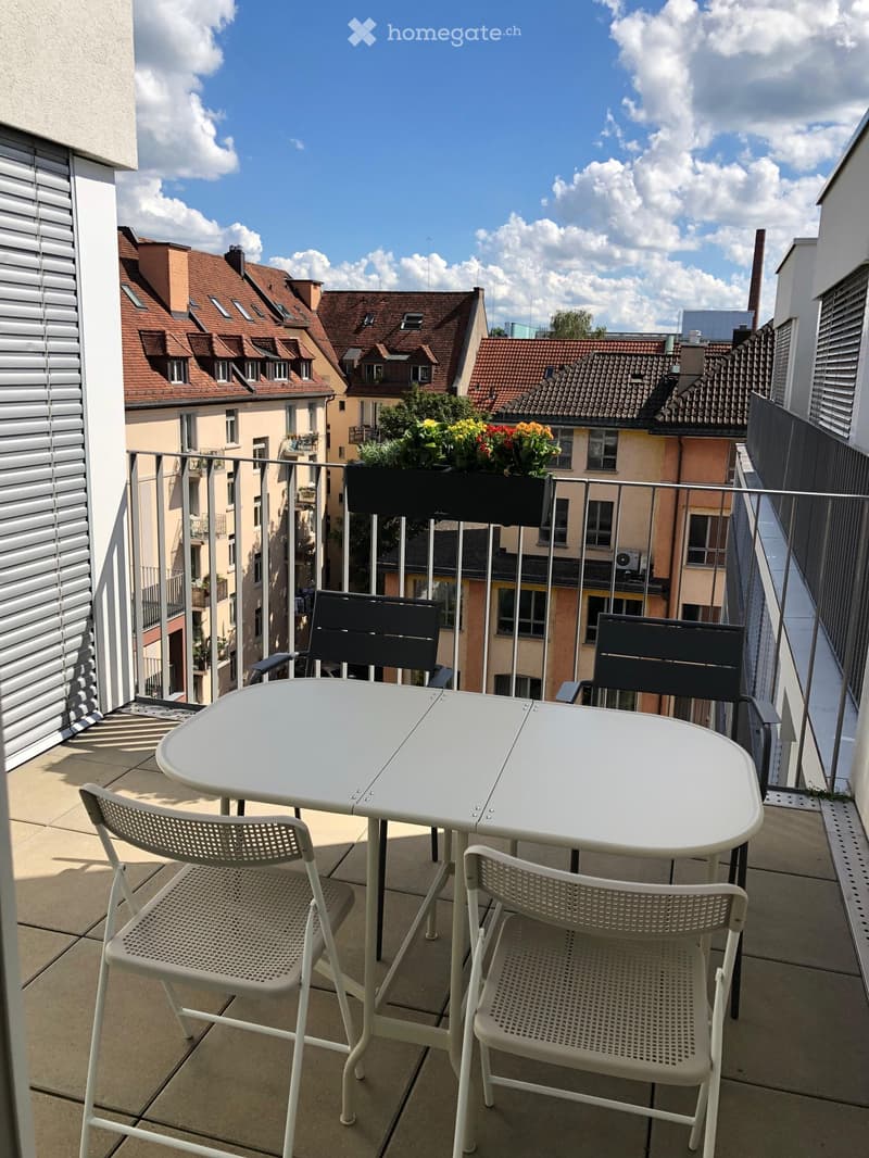 Balcony with a white table and four chairs, overlooking a cityscape with red-roofed buildings and greenery.
