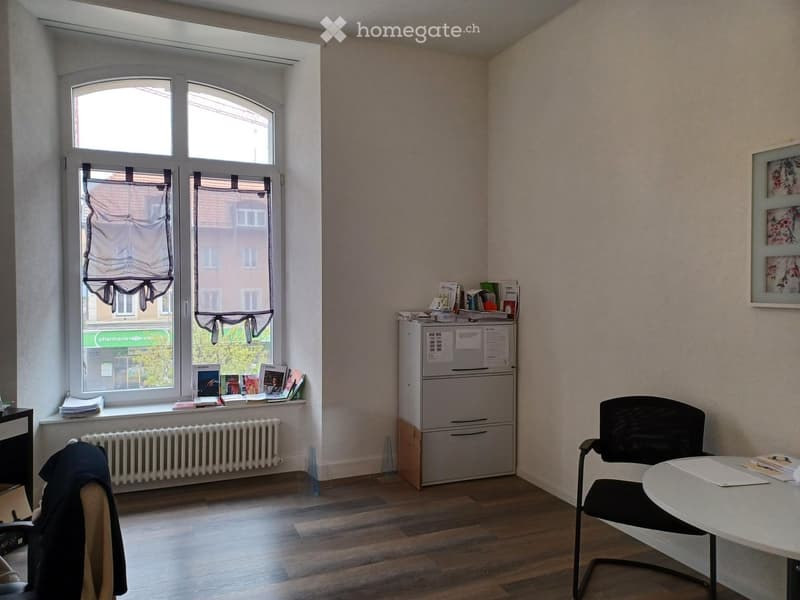 Office space with wooden flooring, large window with blinds, radiator, filing cabinet, chair, table, and books on the windowsill.