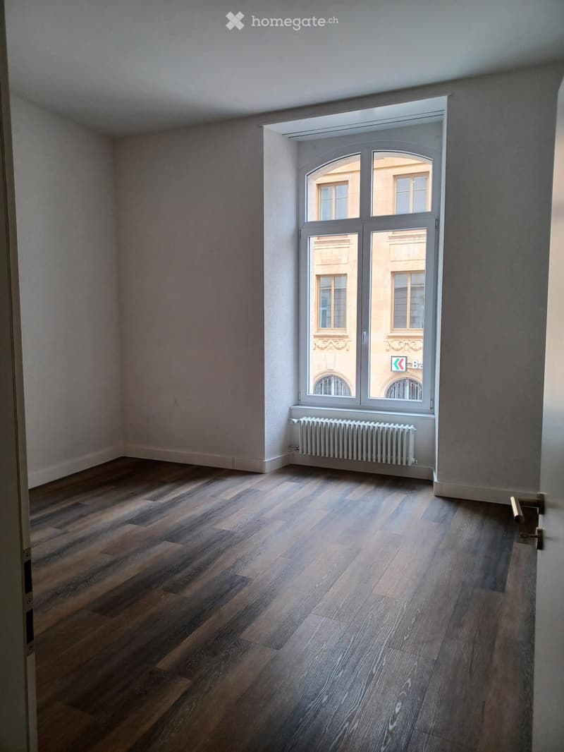 Empty room with wooden flooring, white walls, and a large window with a radiator below it.