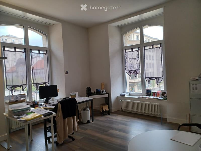Office space with wooden flooring, large windows with curtains, desk with computer, chair, and various office supplies.