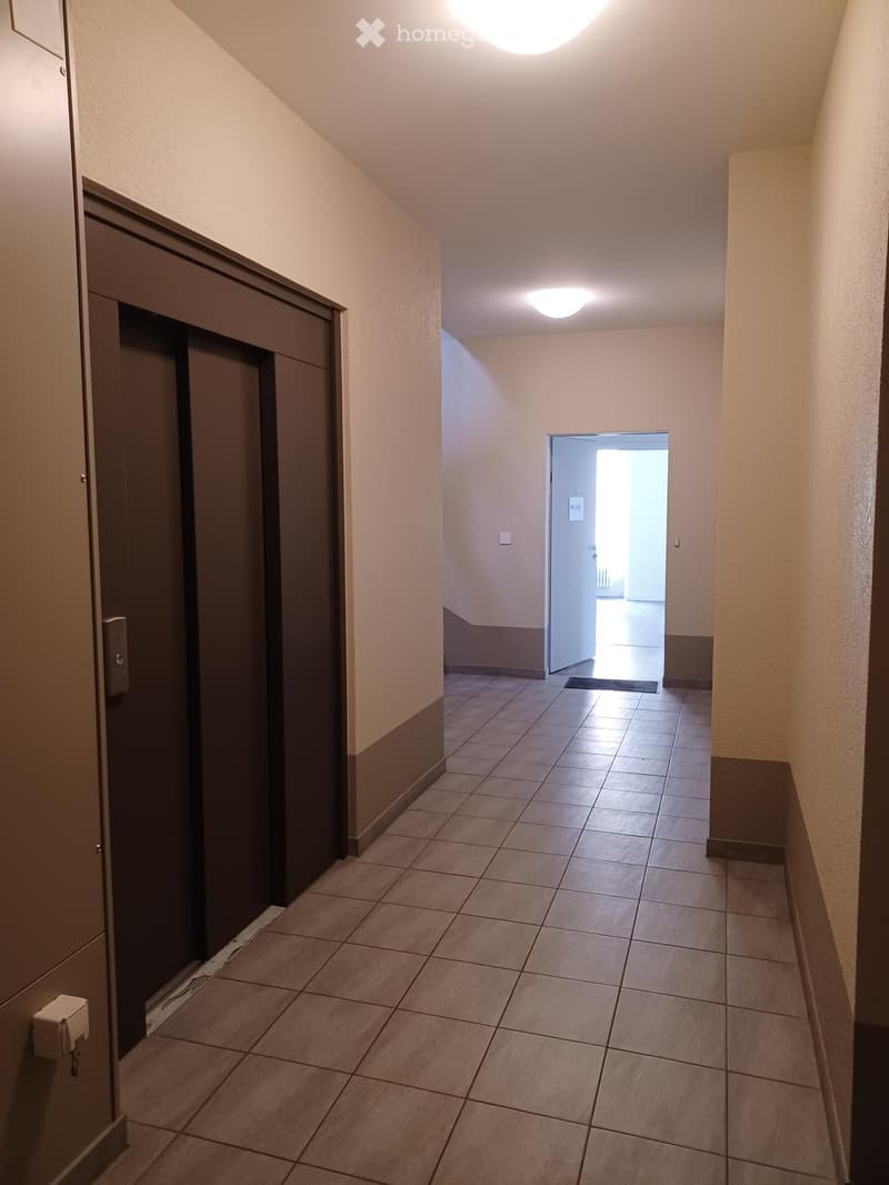 Empty hallway with tiled floor, beige walls, and recessed lighting. Features a closed elevator door on the left and an open doorway leading to another area on the right.