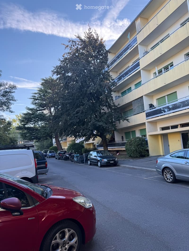 Multi-story residential building with balconies, surrounded by parked cars and trees.
