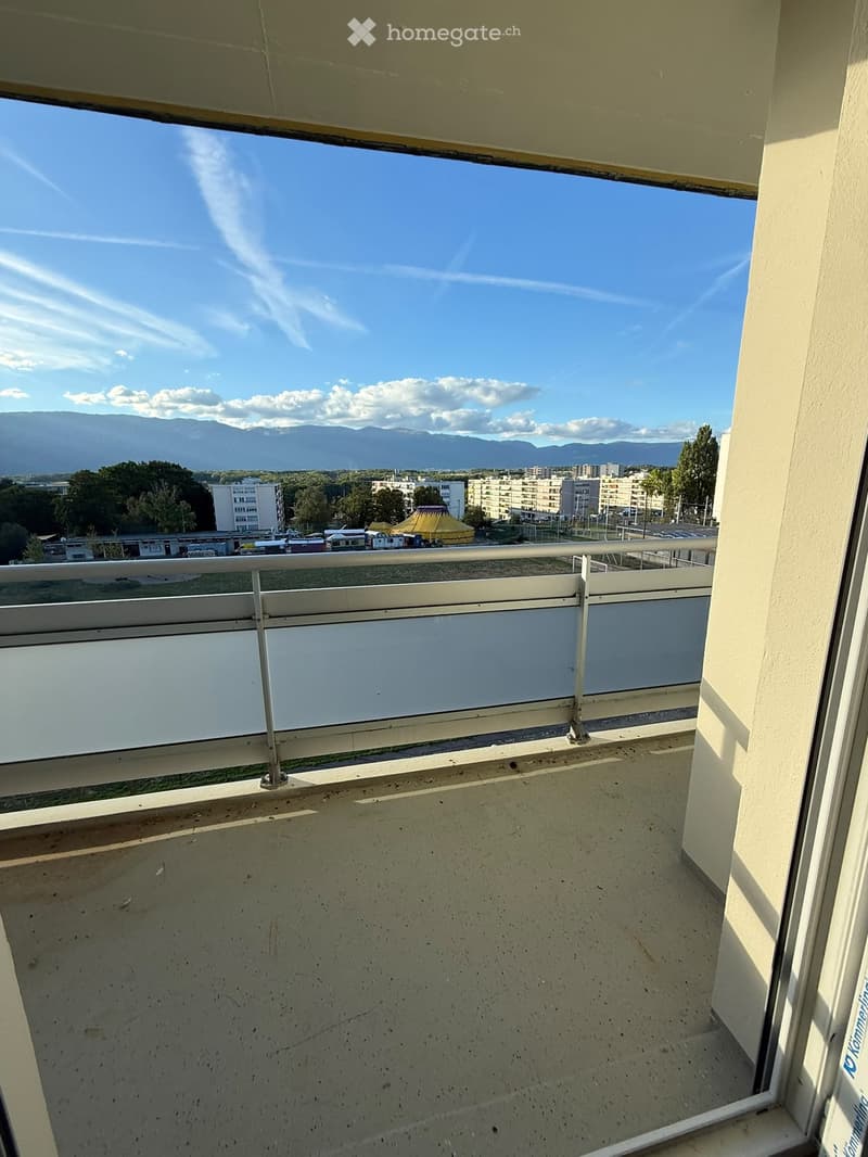 Balcony with metal railing, overlooking a cityscape with buildings, trees, and distant mountains under a clear blue sky.