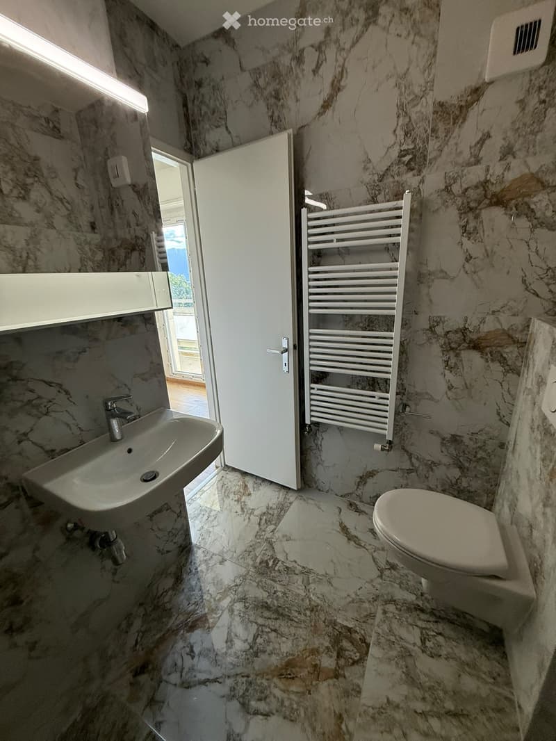 Modern bathroom with marble walls and floor, featuring a sink, toilet, and a white radiator. An open door leads to a balcony area.
