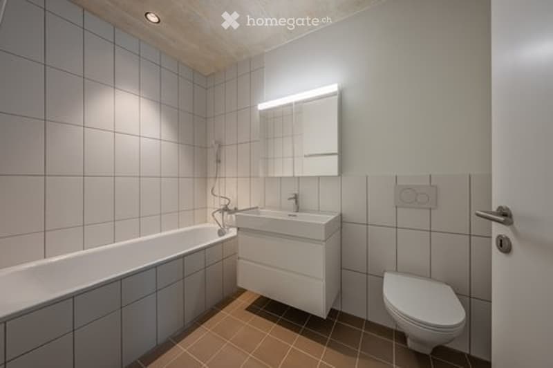 Modern bathroom with white tiles, a bathtub, a sink, a toilet, and a mirror with built-in lighting.