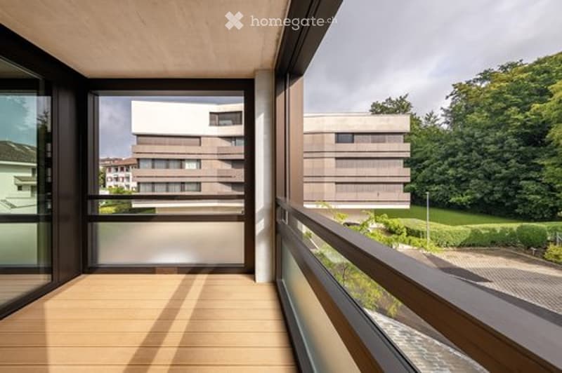 Modern balcony with wooden flooring, glass railings, and a view of a contemporary building and greenery.