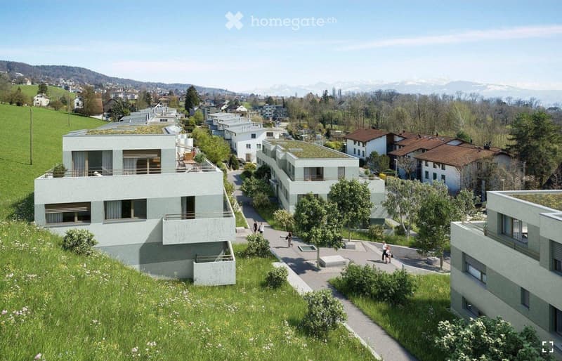 Aerial view of a modern residential complex with multiple buildings, green roofs, and surrounding greenery. The buildings are light-colored with large windows and balconies, situated on a hillside with a scenic backdrop of mountains and trees.