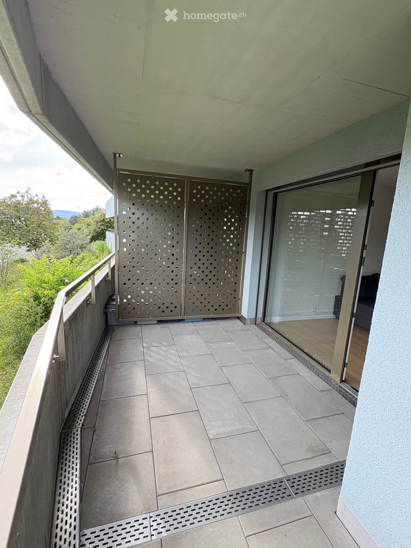 Balcony with tiled floor, metal railing, glass door leading inside, decorative perforated metal panels, and a view of greenery.