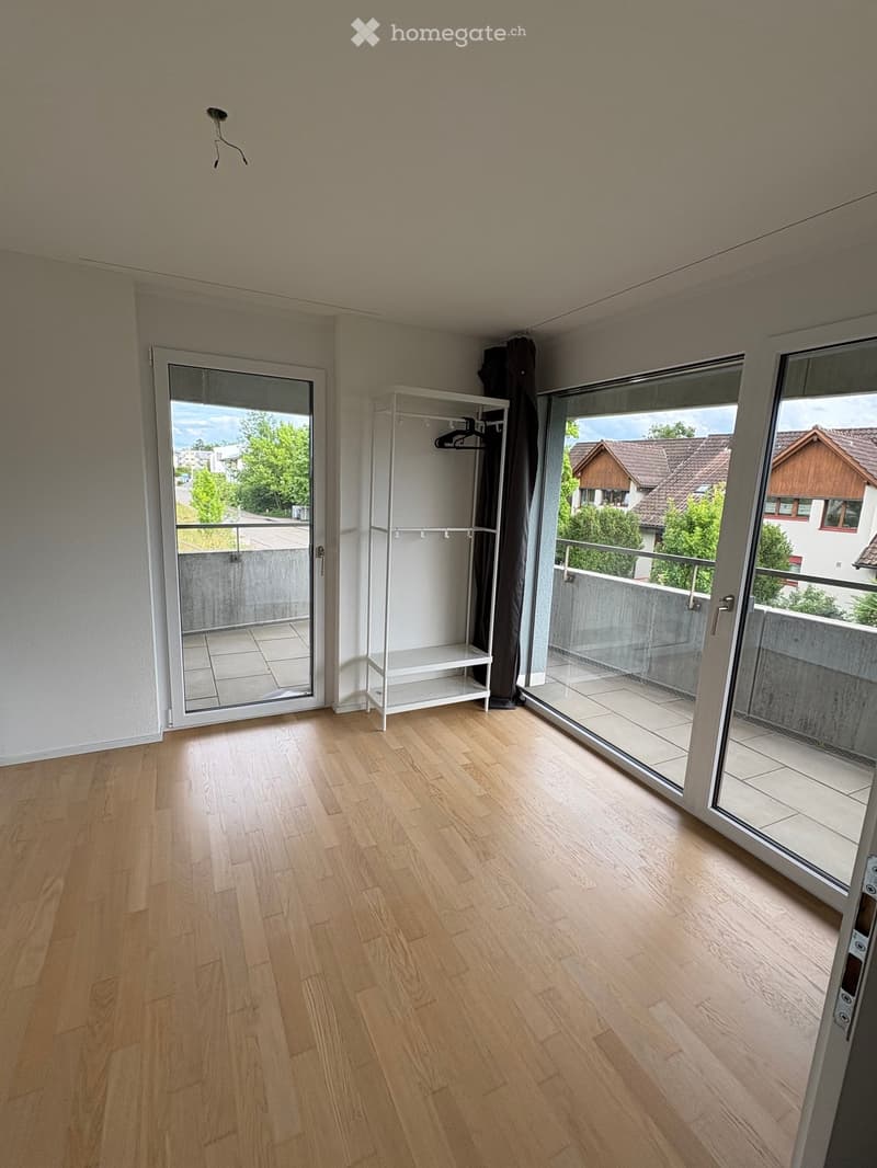 Empty room with wooden flooring, white walls, and large sliding glass doors leading to a balcony with a view of neighboring houses and greenery.