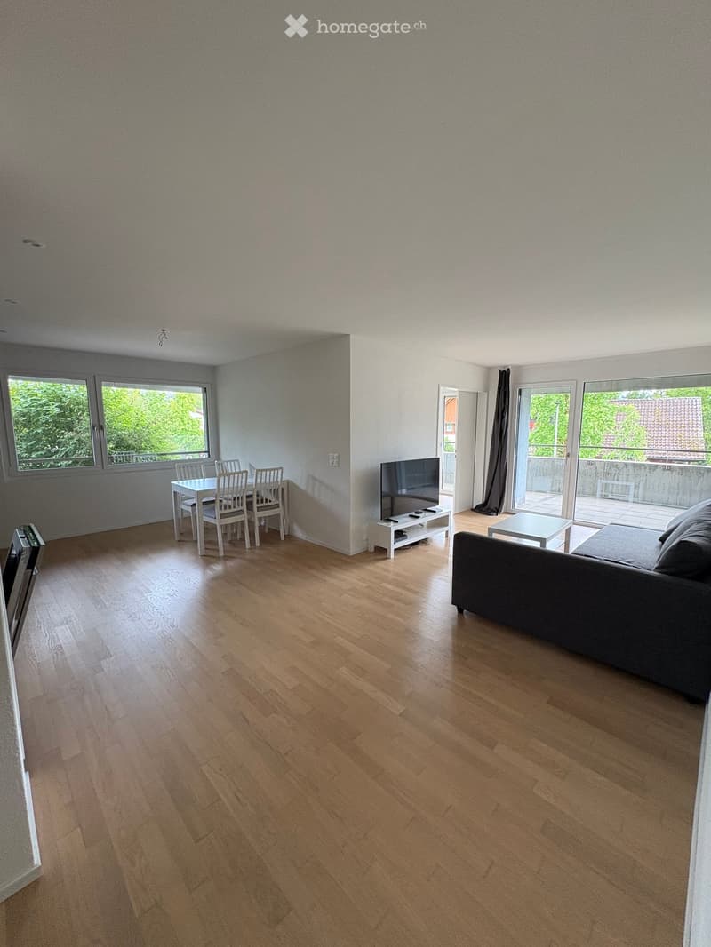 Bright living and dining area with wooden flooring, large windows, and sliding glass doors leading to a balcony. Features a sofa, coffee table, dining table with chairs, and a television.