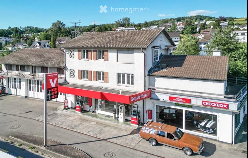 A commercial building with a gas station and car showroom, featuring a red canopy, gas pumps, and a display of vintage cars.