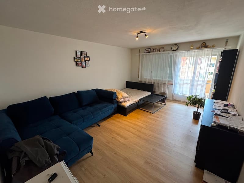A modern living room with a blue sectional sofa, a small black coffee table, a potted plant, and a bed in the corner.