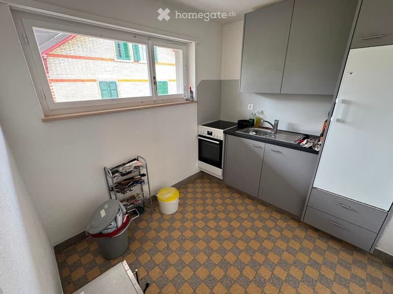 Kitchen with grey cabinets, a sink, oven, refrigerator, and a dish rack. The floor is tiled in a checkered pattern, and there is a window with a view of a neighboring building.