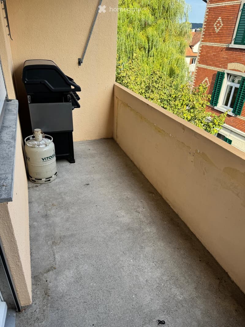 Balcony with concrete floor, gas cylinder, and grill, overlooking greenery and neighboring buildings.