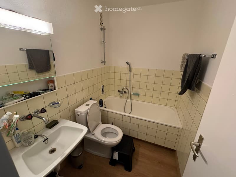 Bathroom with a sink, toilet, bathtub, mirror, and various toiletries. Tiled walls and wooden flooring.
