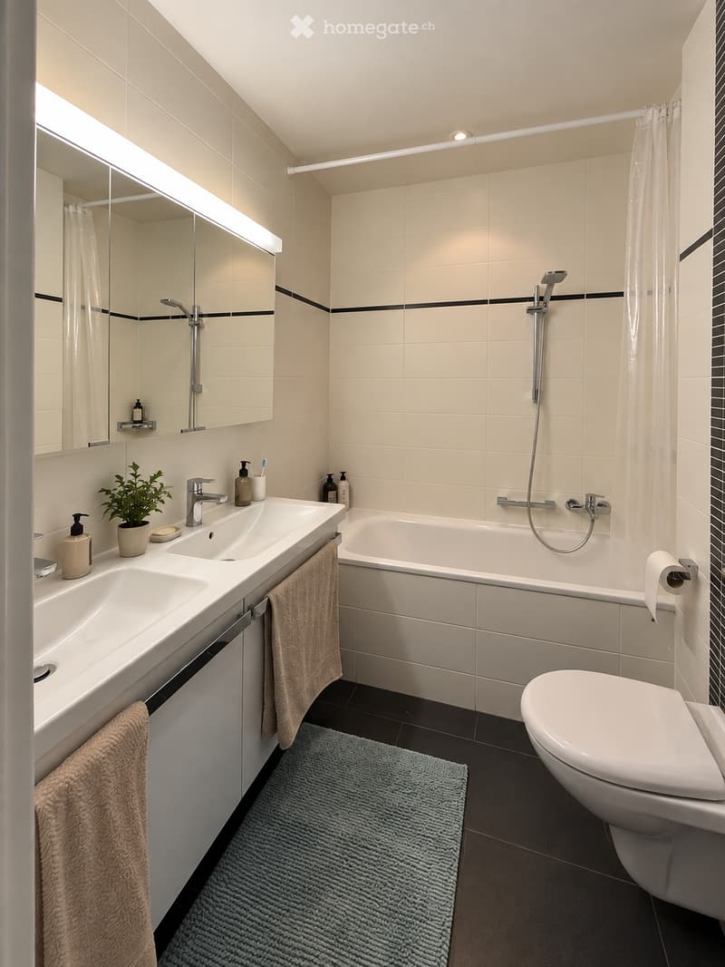 Modern bathroom with a double sink, bathtub, toilet, and a small plant on the sink counter. Features a large mirror with built-in lighting, a showerhead, and a towel hanging on the side.