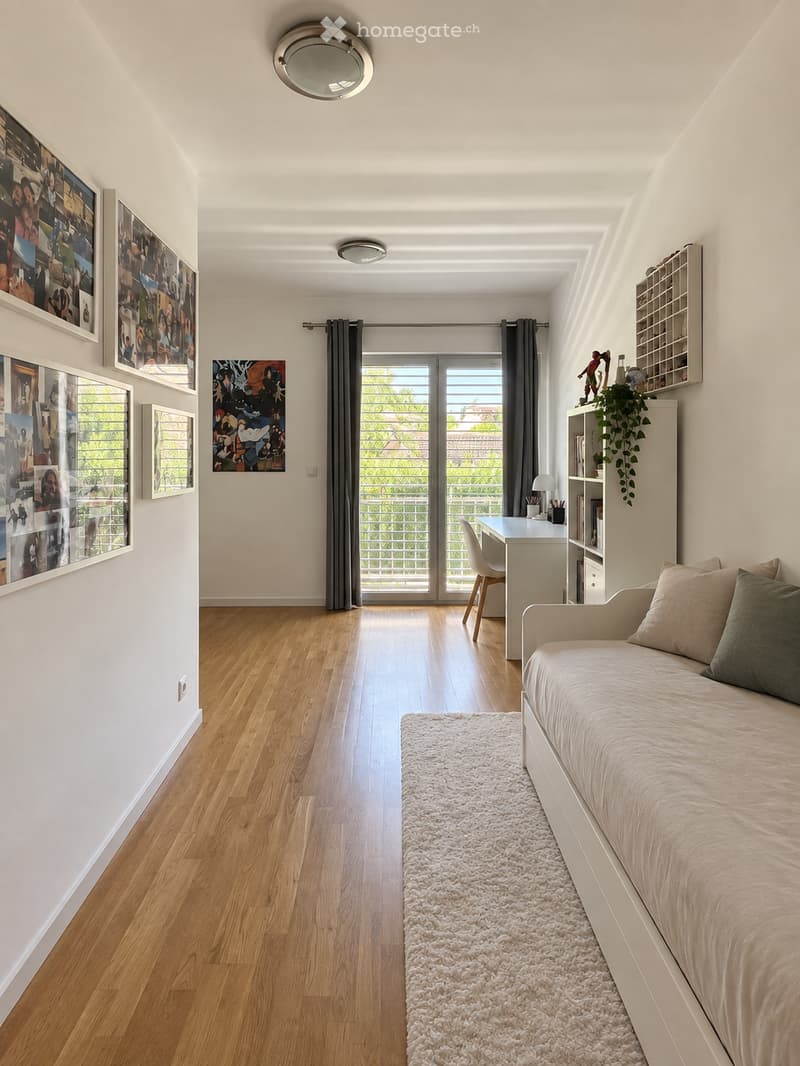 Bright bedroom with wooden flooring, white walls, a daybed with pillows, a fluffy white rug, a desk with a chair, a bookshelf with books and a plant, and several picture frames on the wall.
