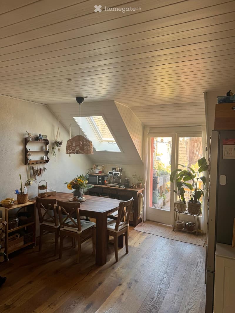 Bright, wooden-floored room with a dining table and chairs, kitchen area with appliances, and large windows with plants and a view.