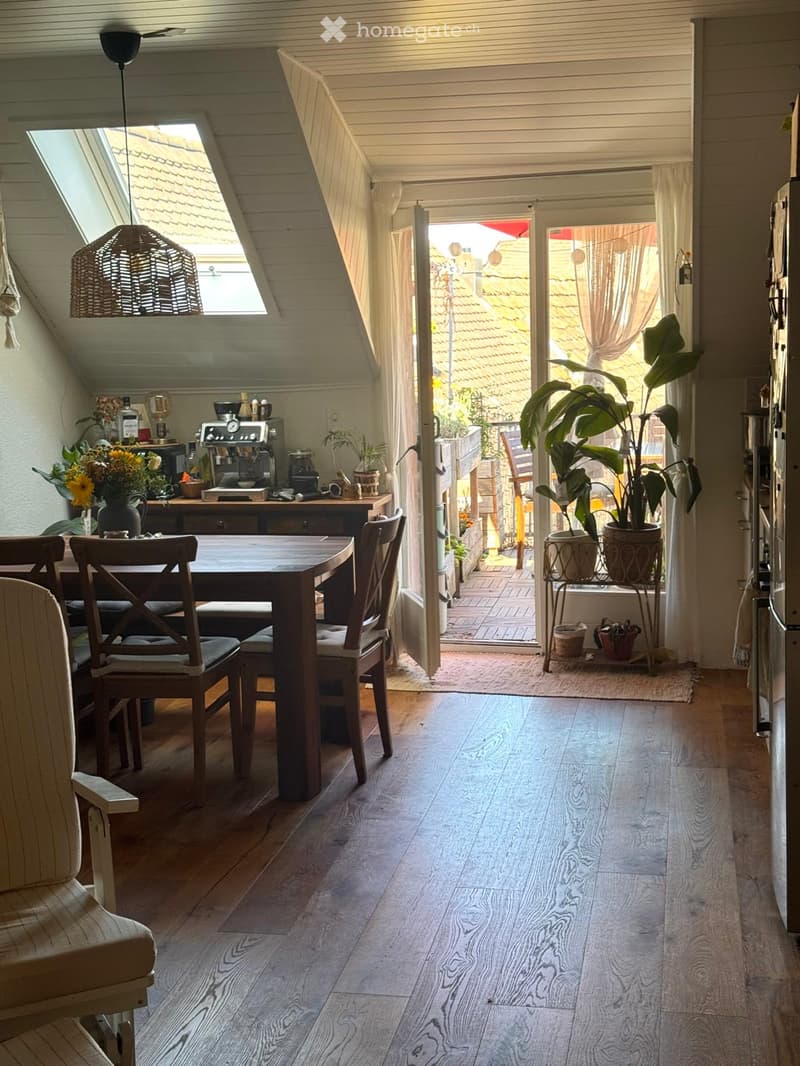 Bright, airy room with wooden flooring, a dining table with chairs, a coffee machine on a counter, and a large potted plant near the balcony entrance.