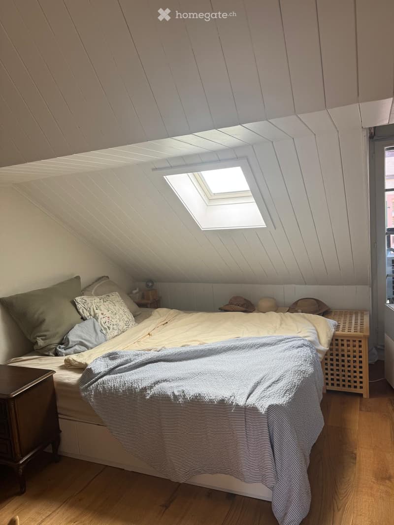 Cozy attic bedroom with slanted wooden ceiling, skylight window, bed with pillows and blanket, nightstand, and decorative hats on a shelf.