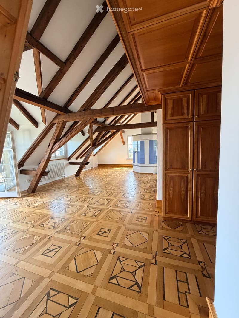 An attic space with exposed wooden beams, a patterned wooden floor, and white walls. The room features wooden doors and windows, with natural light streaming in from the windows.