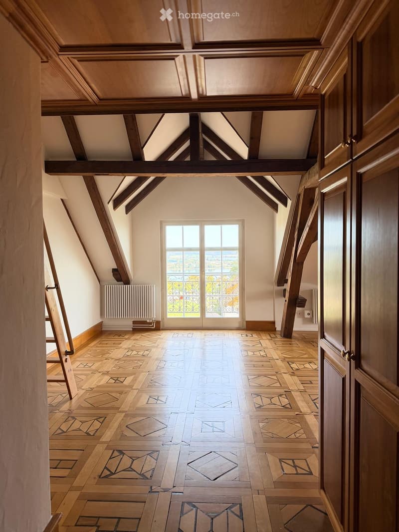 An empty attic space with exposed wooden beams, a large window with decorative ironwork, and a patterned wooden floor.