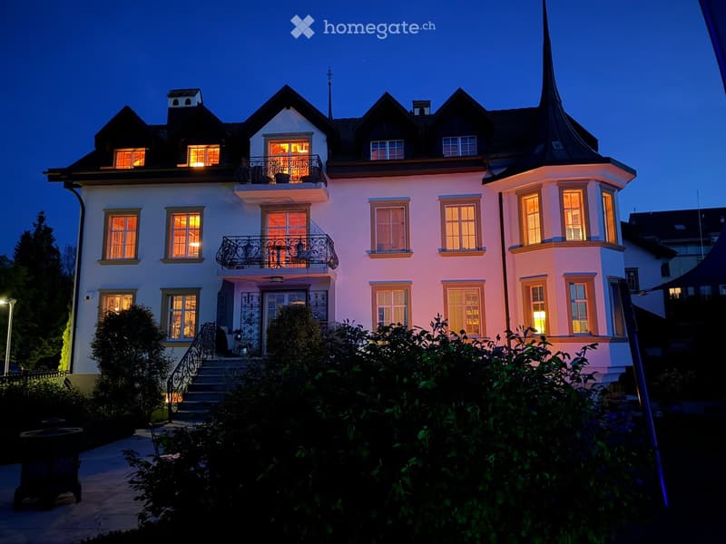 Large, elegant two-story house with white walls, multiple windows, and a steeply pitched roof featuring dormers and a prominent spire. The house is illuminated from within, with warm lighting visible through the windows, and is surrounded by well-maintain