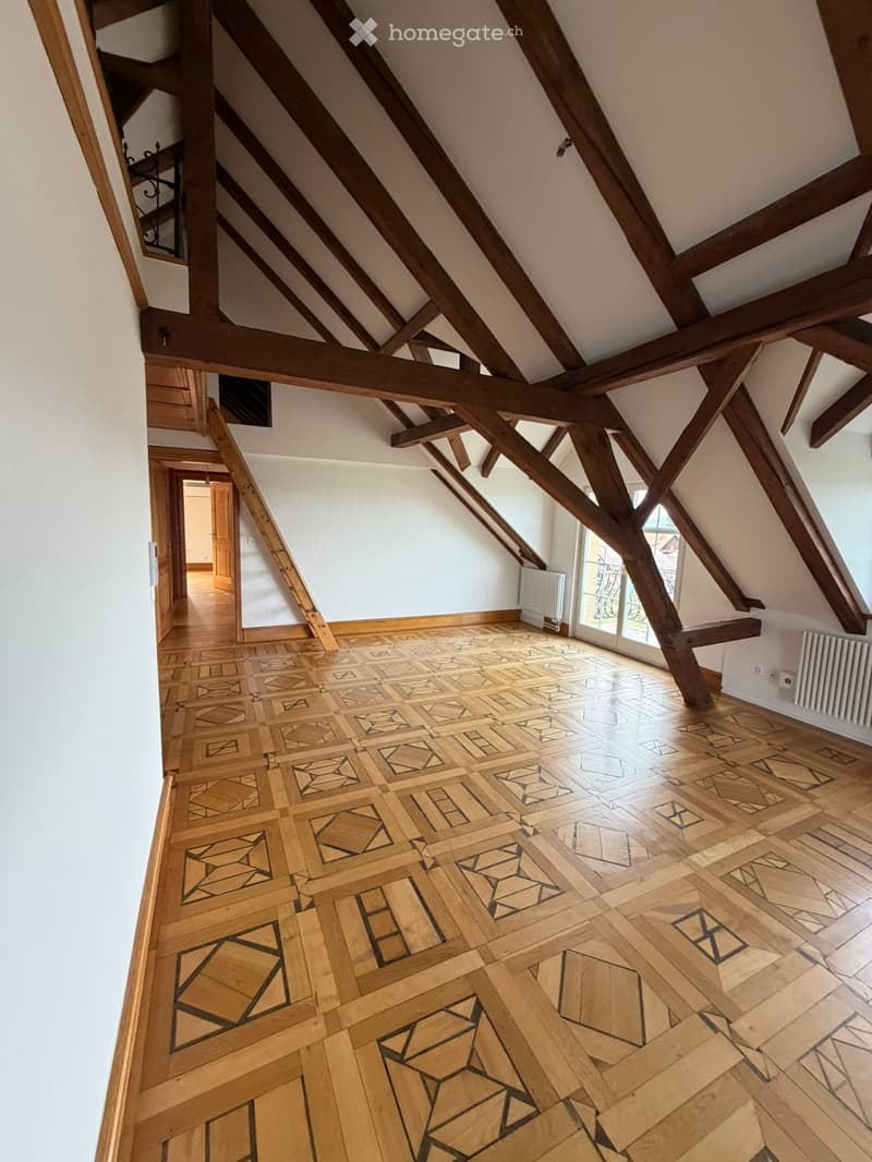 An attic space with exposed wooden beams, a patterned wooden floor, and large windows allowing natural light to enter. The room is empty, with no furniture or decorations visible.