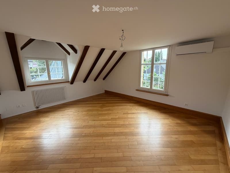 Empty attic room with wooden flooring, white walls, exposed wooden beams, and large windows allowing natural light.
