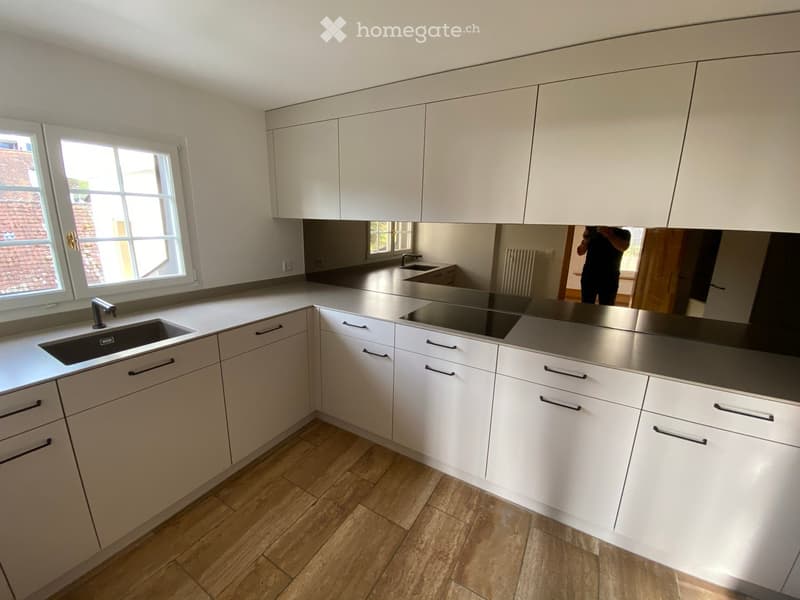 Modern kitchen with white cabinets, stainless steel countertops, large windows, wooden flooring, single sink, black induction stove, mirror with reflection, small room at back