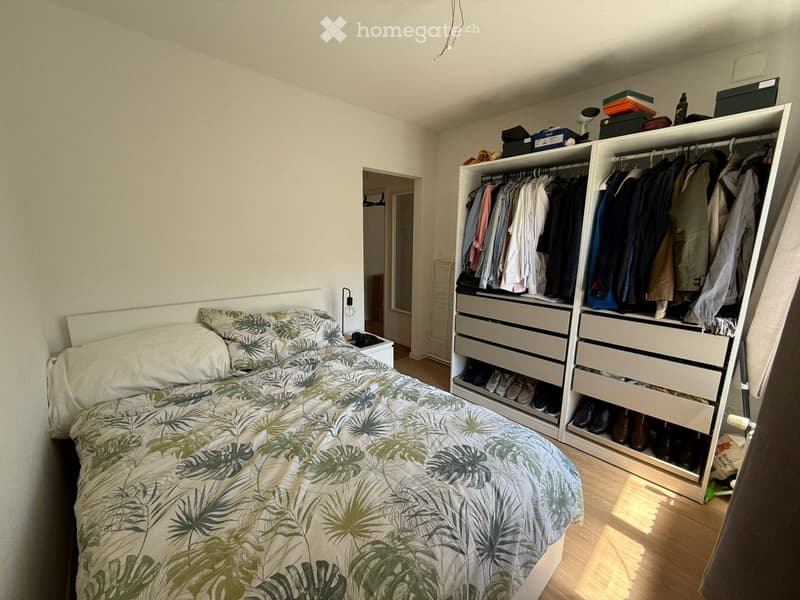 Bedroom with a bed covered in a palm leaf patterned duvet, a large wardrobe with hanging clothes and drawers, and a nightstand with a lamp.