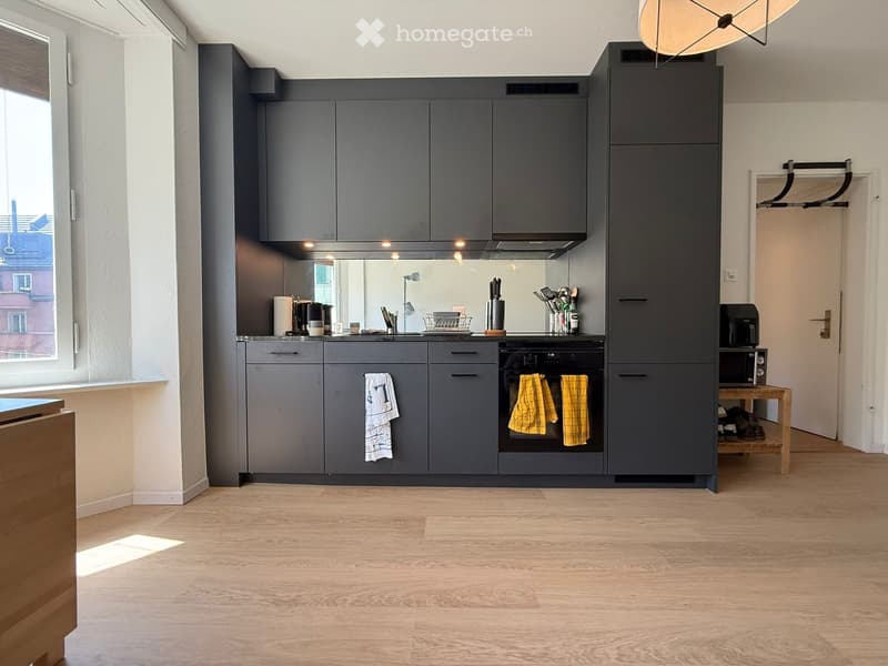 Modern kitchen with grey cabinets, wooden flooring, and various appliances including an oven, microwave, and sink. Towels are hanging from the oven handle and a knife block is on the counter.
