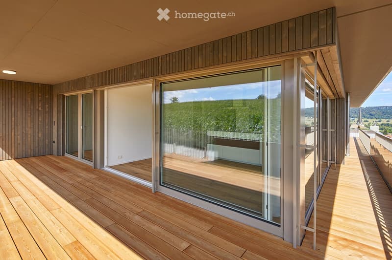 Modern balcony with large glass sliding doors, wooden decking, and a view of greenery and hills.
