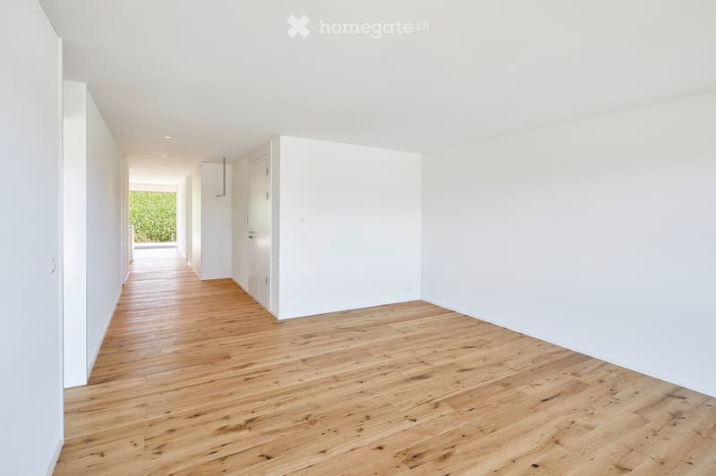 Empty hallway with light wooden flooring and white walls, featuring a door at the end and a view of greenery outside.