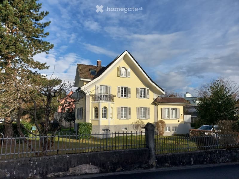 Two-story yellow house with white shutters, multiple windows, and a small balcony on the upper floor.