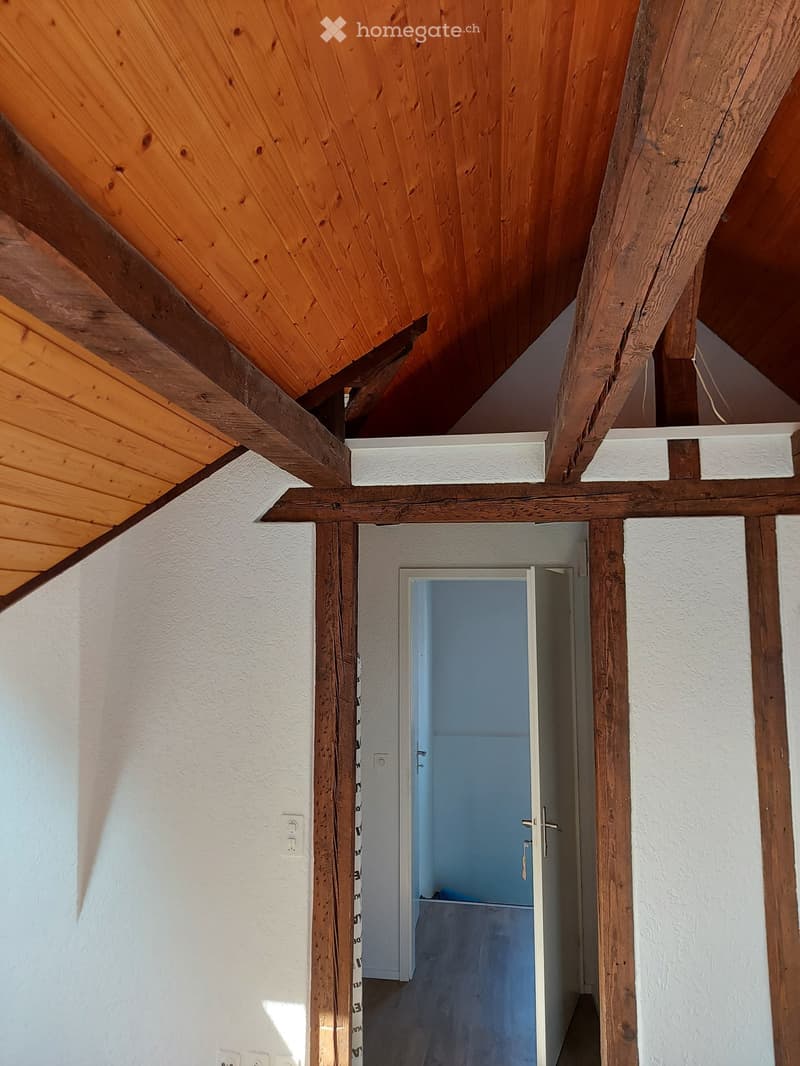 A narrow hallway with wooden beams and white walls, leading to an open doorway with a blue interior visible beyond.