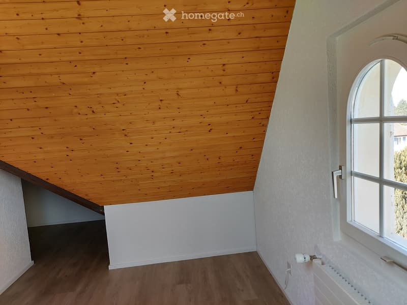 Empty attic room with wooden ceiling, wooden floor, white walls, and a large arched window letting in natural light.