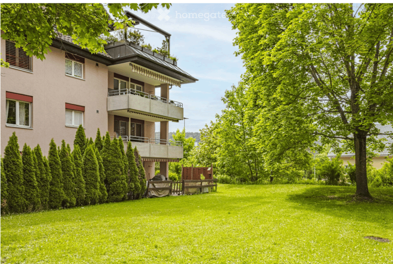 A two-story residential building with a balcony, surrounded by a well-maintained lawn and trees.