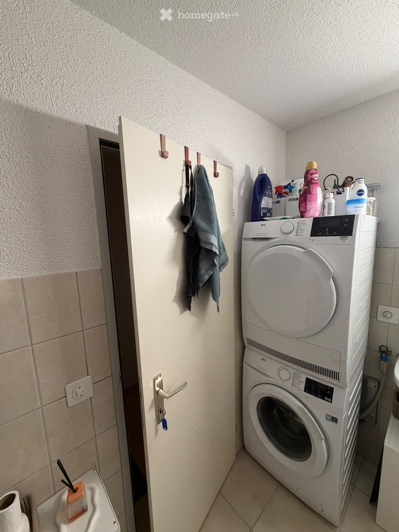 Bathroom with a stacked washing machine and dryer, a towel hanging on a rack, cleaning supplies on top of the dryer, and a partially open door leading to another room.