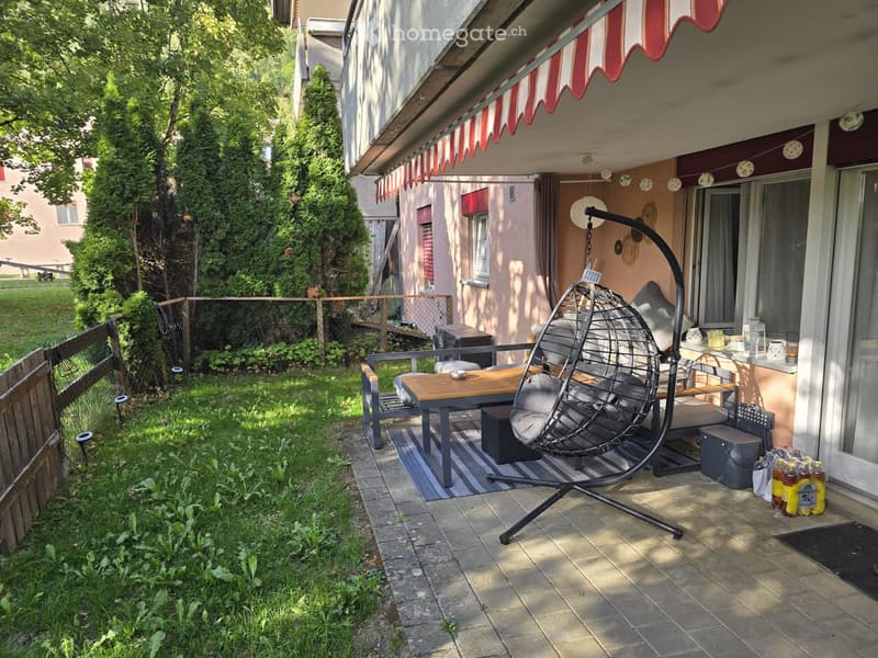 Outdoor patio area with a wooden table, chairs, a hanging wicker chair, a bench, and decorative items including a lantern and cups.