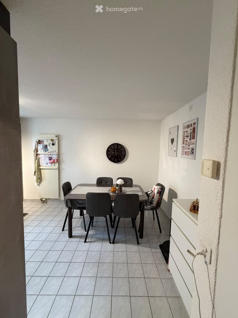 Dining room with a tiled floor, a dining table surrounded by six chairs, a wall-mounted clock, a refrigerator with magnets, and a small cabinet.