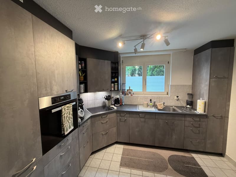 Modern kitchen with grey cabinets, stainless steel appliances, and a window overlooking a green area. The kitchen features an oven, sink, and various small appliances on the counter.