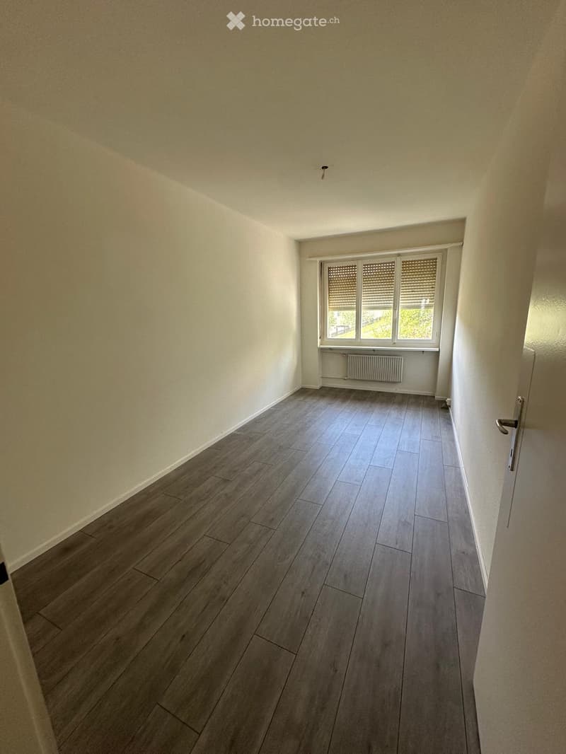 Empty hallway with wooden flooring, white walls, and a window with blinds on the right side.