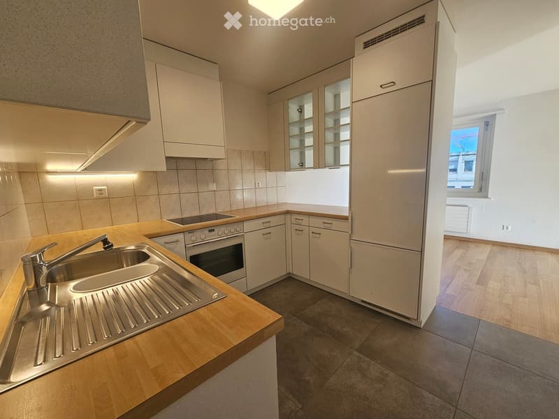 Modern kitchen with light wooden cabinets, stainless steel sink, oven, and countertop. The space is well-lit with recessed lighting and has a window providing natural light.