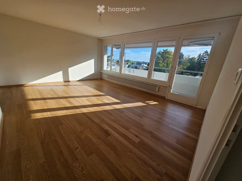 Empty room with wooden flooring and large windows offering a view of the city and trees. The room is unfurnished with natural light streaming in.