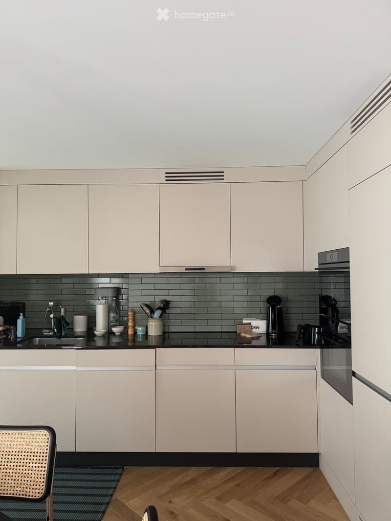 Modern kitchen with light grey cabinets, dark grey tiled backsplash, and wooden flooring. The countertop features a sink, coffee maker, and various kitchen items.